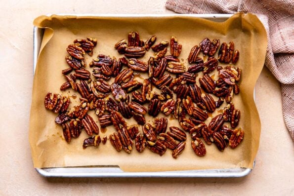 Maple pecans for make ahead sweet potato casserole rest atop a parchment lined baking sheet that sits atop a light peach colored textured surface. A muted red linen napkin rests alongside the baking sheet.