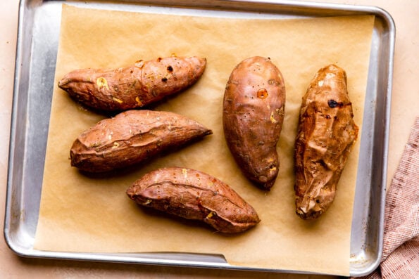 5 large baked sweet potatoes rest atop a parchment lined baking sheet. The baking sheet sits atop a light peach colored textured surface. A muted red linen napkin rests alongside the baking sheet pan.