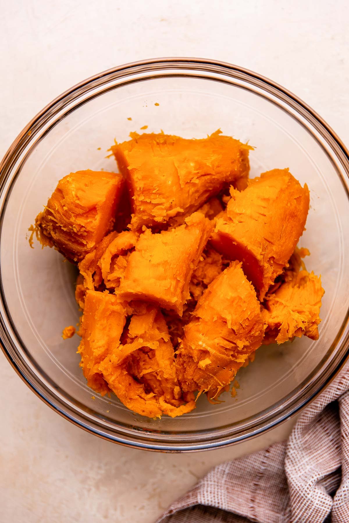 Baked and peeled sweet potatoes fill a large glass mixing bowl that sits atop a light peach colored textured surface. A muted red linen napkin rests alongside the bowl.