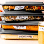 A stack of four glass casserole dishes with gray lids are filled with and labeled as make ahead mashed potatoes, make ahead sweet potato casserole, make ahead green casserole, and make ahead stuffing. A mason jar filled with and labeled as make ahead gravy rests is placed in front of the casserole storage dishes. The dishes sit atop a light peach textured surface with a peach background. A red muted linen napkin and Fall colored florals rest in the background.