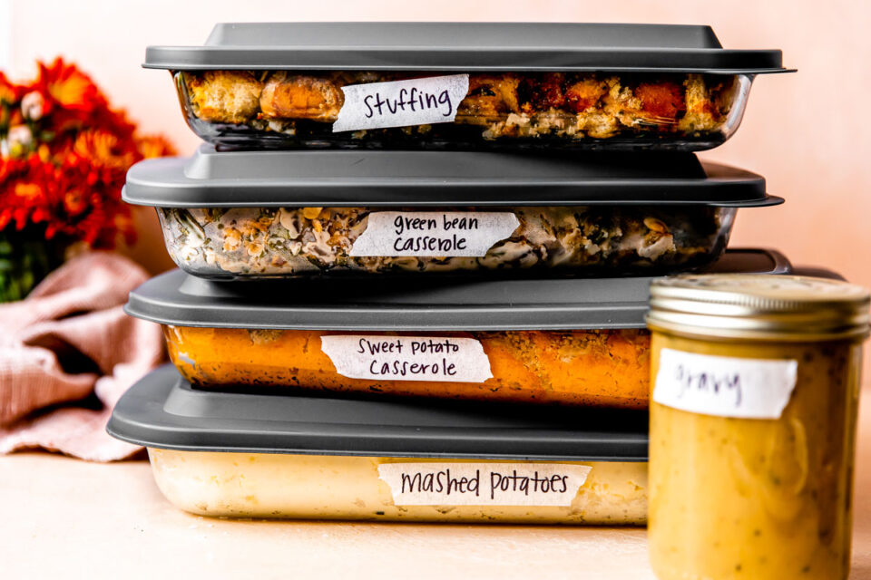 A stack of four glass casserole dishes with gray lids are filled with and labeled as make ahead mashed potatoes, make ahead sweet potato casserole, make ahead green casserole, and make ahead stuffing. A mason jar filled with and labeled as make ahead gravy rests is placed in front of the casserole storage dishes. The dishes sit atop a light peach textured surface with a peach background. A red muted linen napkin and Fall colored florals rest in the background.