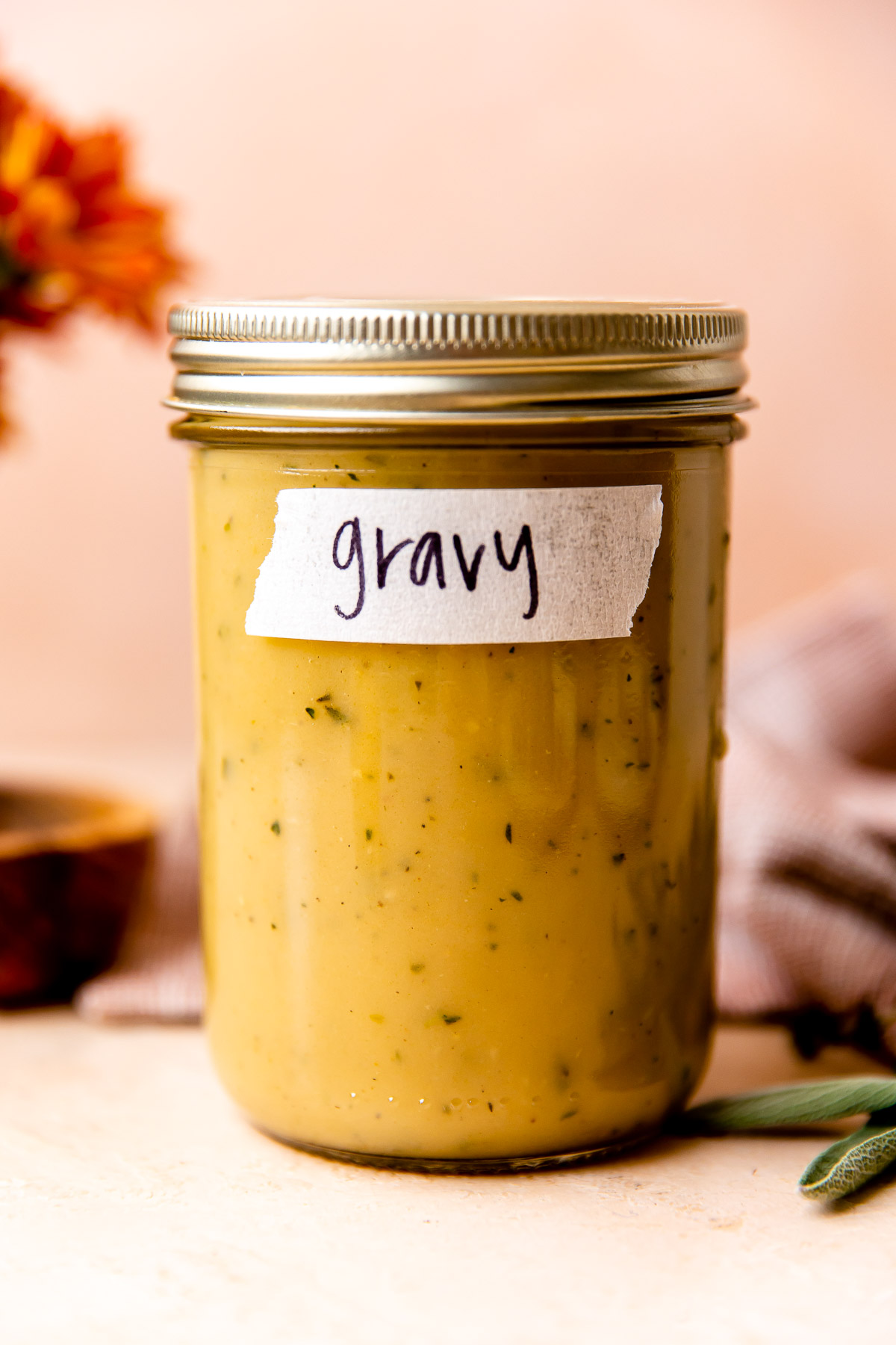 Make ahead turkey gravy fills a large clear glass mason jar. The jar sits atop a light peach colored textured surface and is surrounded by orange florals, a small wooden pinch bowl filled with kosher salt, a muted red linen napkin, and loose sprigs of fresh herbs.