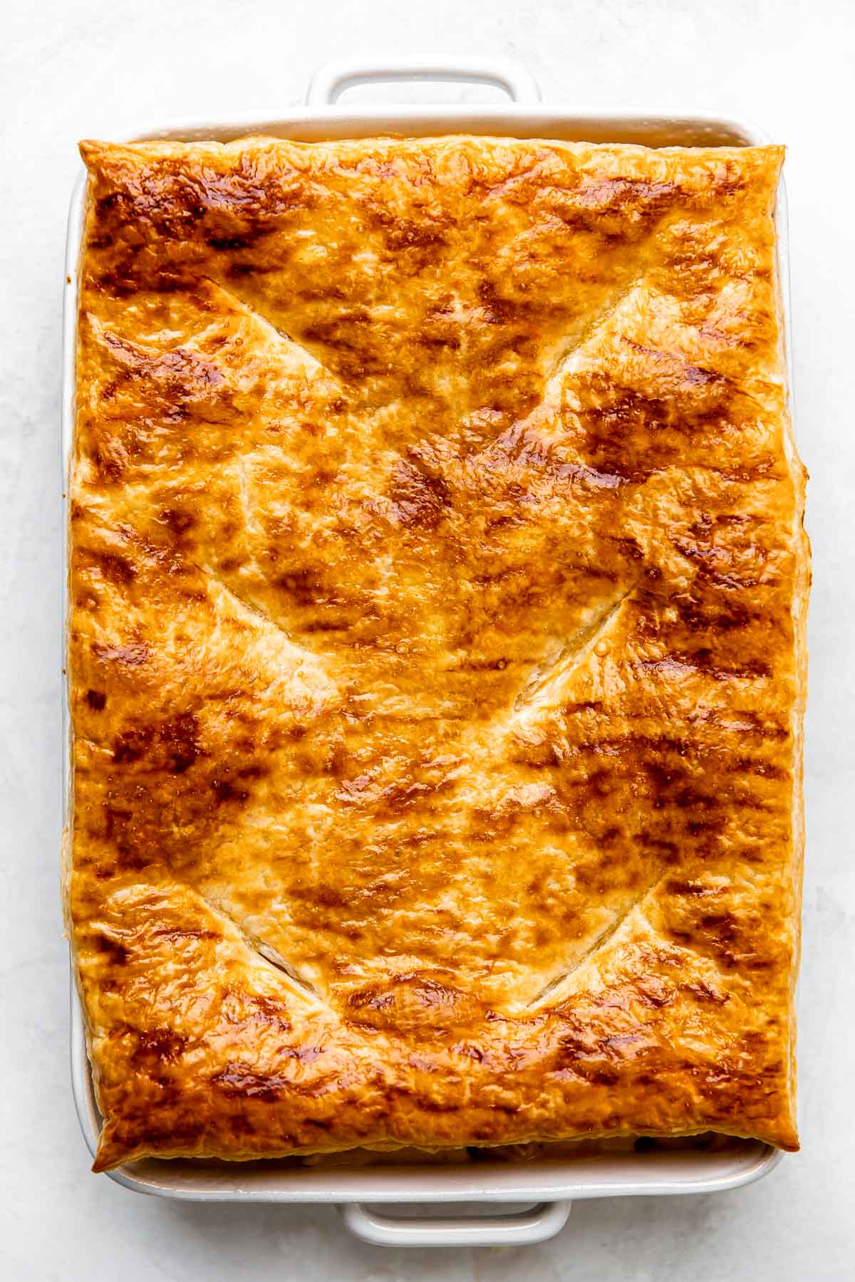 A par-baked sheet of puff pastry is placed overtop of an assembled seafood pot pie inside of a large white baking dish. The baking dish sits atop of a creamy white textured surface.