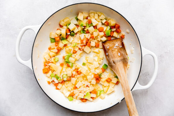 Diced carrot, yellow onion, celery, and Russet potato fill a large white double-handled braising pan. The pan sits atop a creamy white textured surface with a wooden spatula resting inside for stirring.