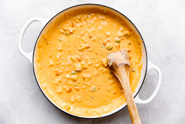 Seafood pot pie sauce fills a large white double-handled pan that sits atop a creamy white textured surface. A wooden spatula rests inside of the pan for stirring.