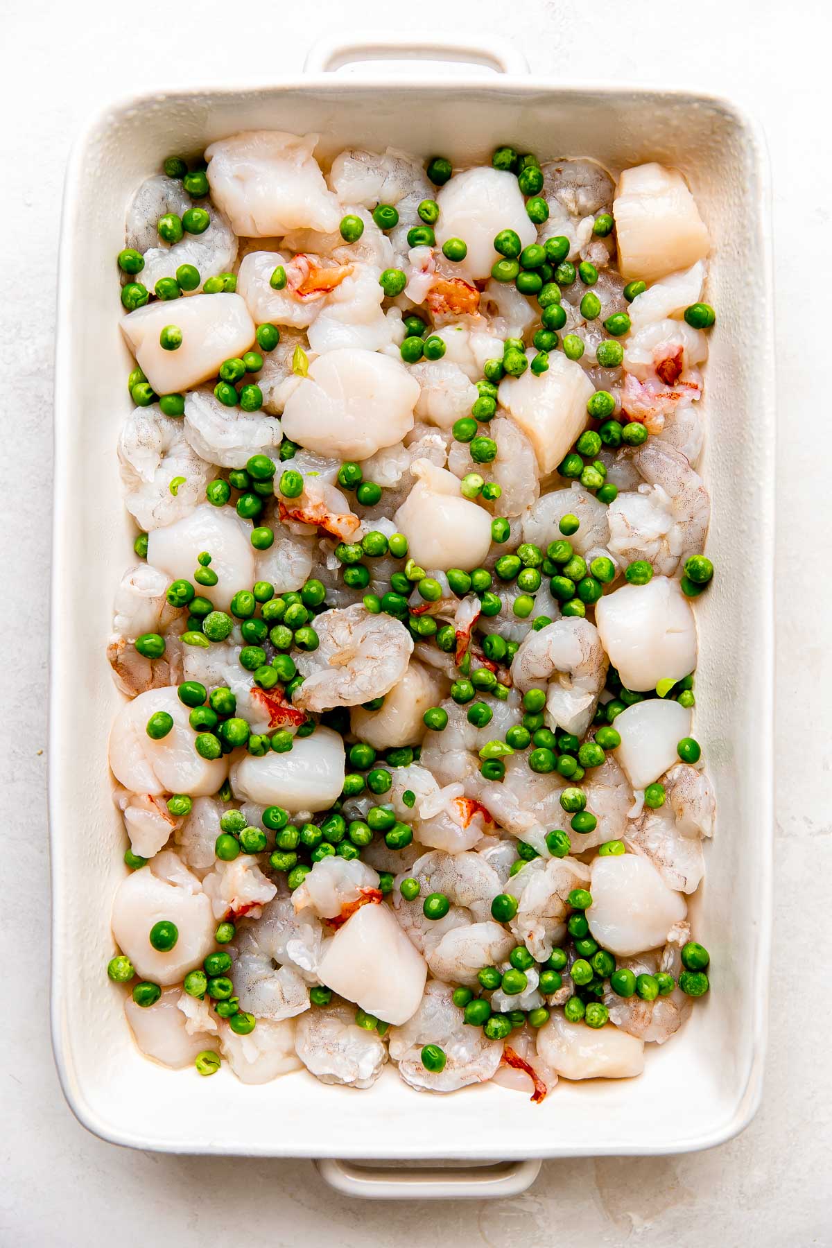 A layer of scallops, shrimp, lobster, & peas fills a white oven-safe baking dish that sits atop a creamy white textured surface. This is the first layer of a partially assembled seafood pot pie.