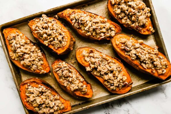 Eight assembled twice baked sweet potatoes with streusel topping are arranged atop a parchment lined baking sheet. The sheet pan sits atop a white and gray marble surface.