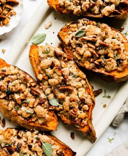 A close up shot of five twice baked sweet potatoes with streusel topping are arranged atop a white oval serving platter. The platter sits atop a white and gray marble surface surrounded a small white scalloped pinch bowl filled with chopped pecans and walnuts, loose sprigs of fresh herbs, and a light cream colored linen napkin.