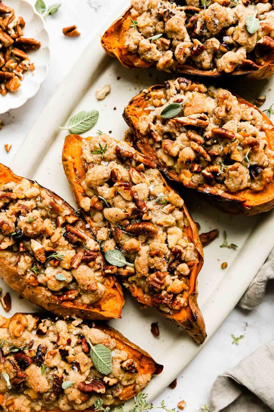 A close up shot of five twice baked sweet potatoes with streusel topping are arranged atop a white oval serving platter. The platter sits atop a white and gray marble surface surrounded a small white scalloped pinch bowl filled with chopped pecans and walnuts, loose sprigs of fresh herbs, and a light cream colored linen napkin.