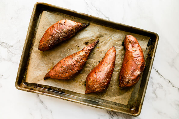 Four medium baked sweet potatoes are arranged atop a parchment lined baking sheet pan that sits atop a white and gray marble surface.