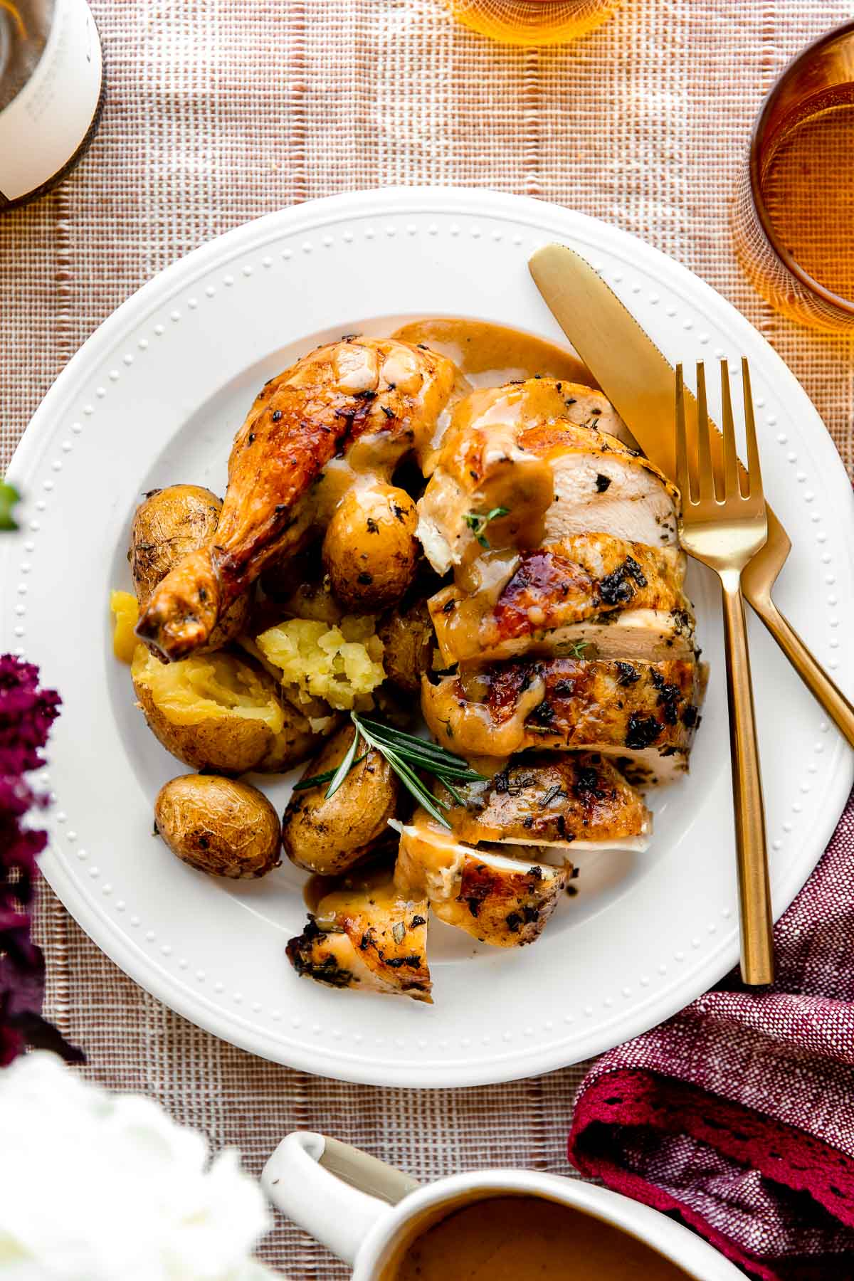An overhead shot of Thanksgiving chicken served atop a white dinner plate that sits atop a muted red table cloth with a gold fork & knife resting atop of the plate. The oven roasted chicken is served with potatoes, chicken gravy and is garnished with fresh herbs. The plate is surrounded by a bottle of wine, purple florals, a purple linen napkin, two amber colored drinking glass, and a white gravy boat filled with chicken gravy.