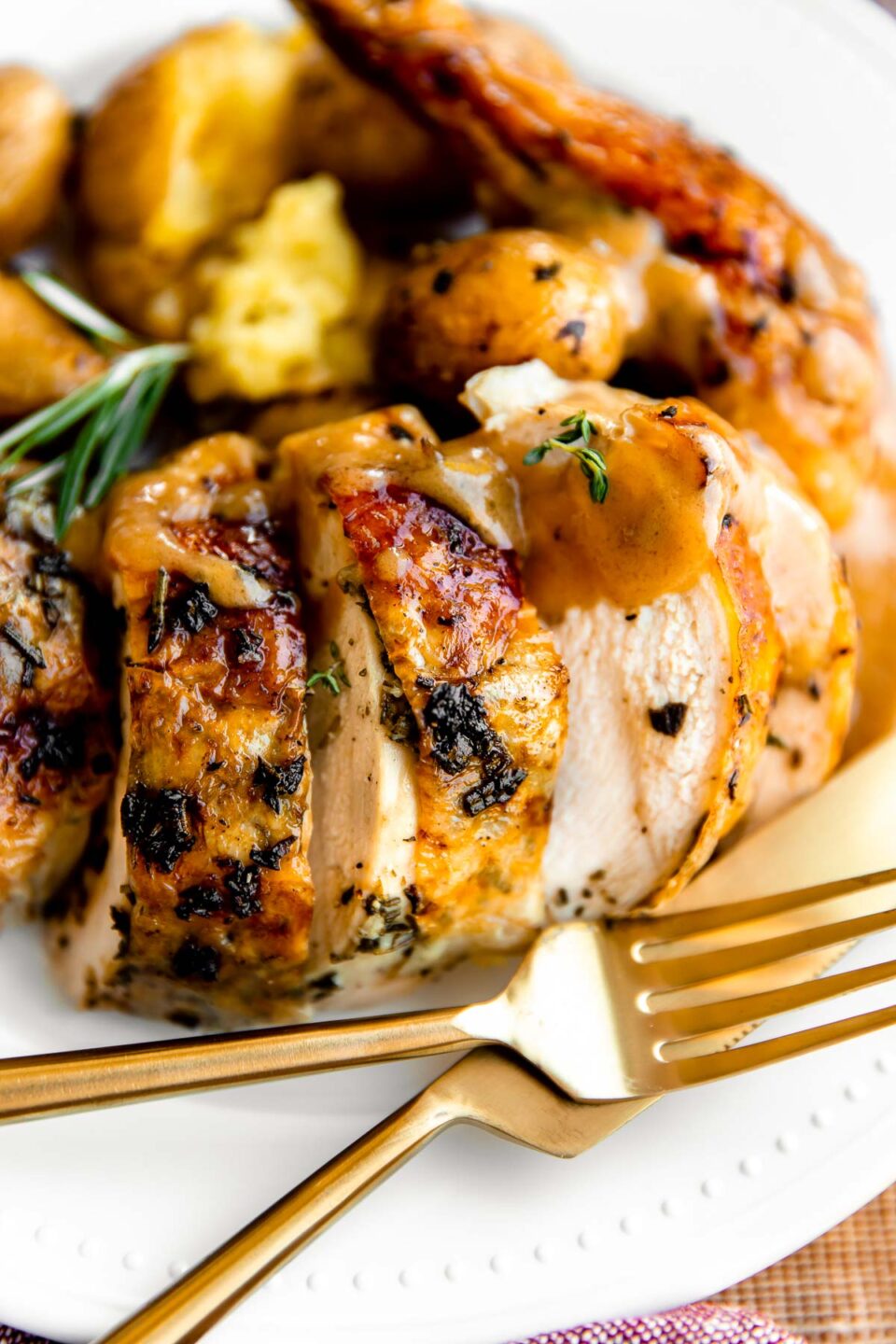 A close up of Thanksgiving chicken served on a white dinner plate with gold silverware. The oven roasted whol chicken has been served with potatoes, chicken gravy, and garnished with fresh herbs.