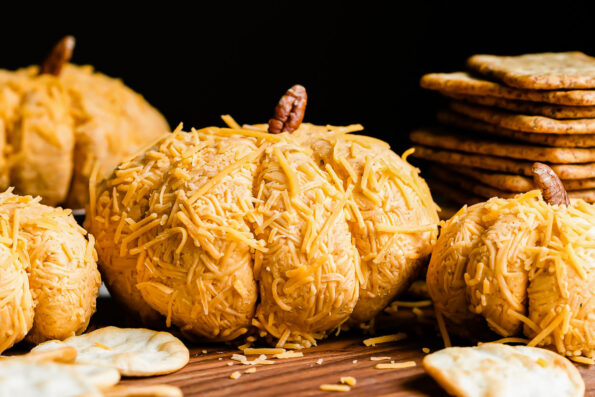 Various sizes of pumpkin-shaped cheese balls are arranged atop a wood serving platter to create a thanksgiving cheese ball pumpkin patch. The platter is surrounded by stacks of crackers and pita chips and sits atop a gray textured surface.