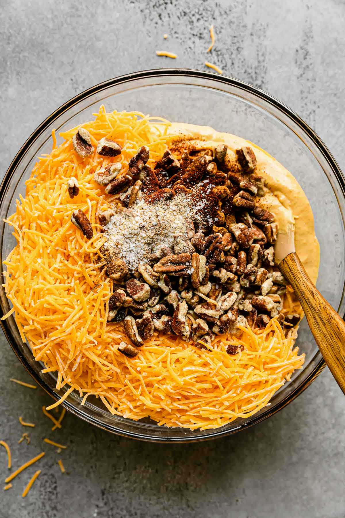 Cream cheese and pumpkin mixture fill a large glass mixing bowl that sits atop a gray textured surface. Shredded cheese, spices, and chopped pecans and walnuts rest atop the cream cheese & pumpkin mixture with a spatula resting inside of the bowl to mix into a pumpkin cheese ball mixture.
