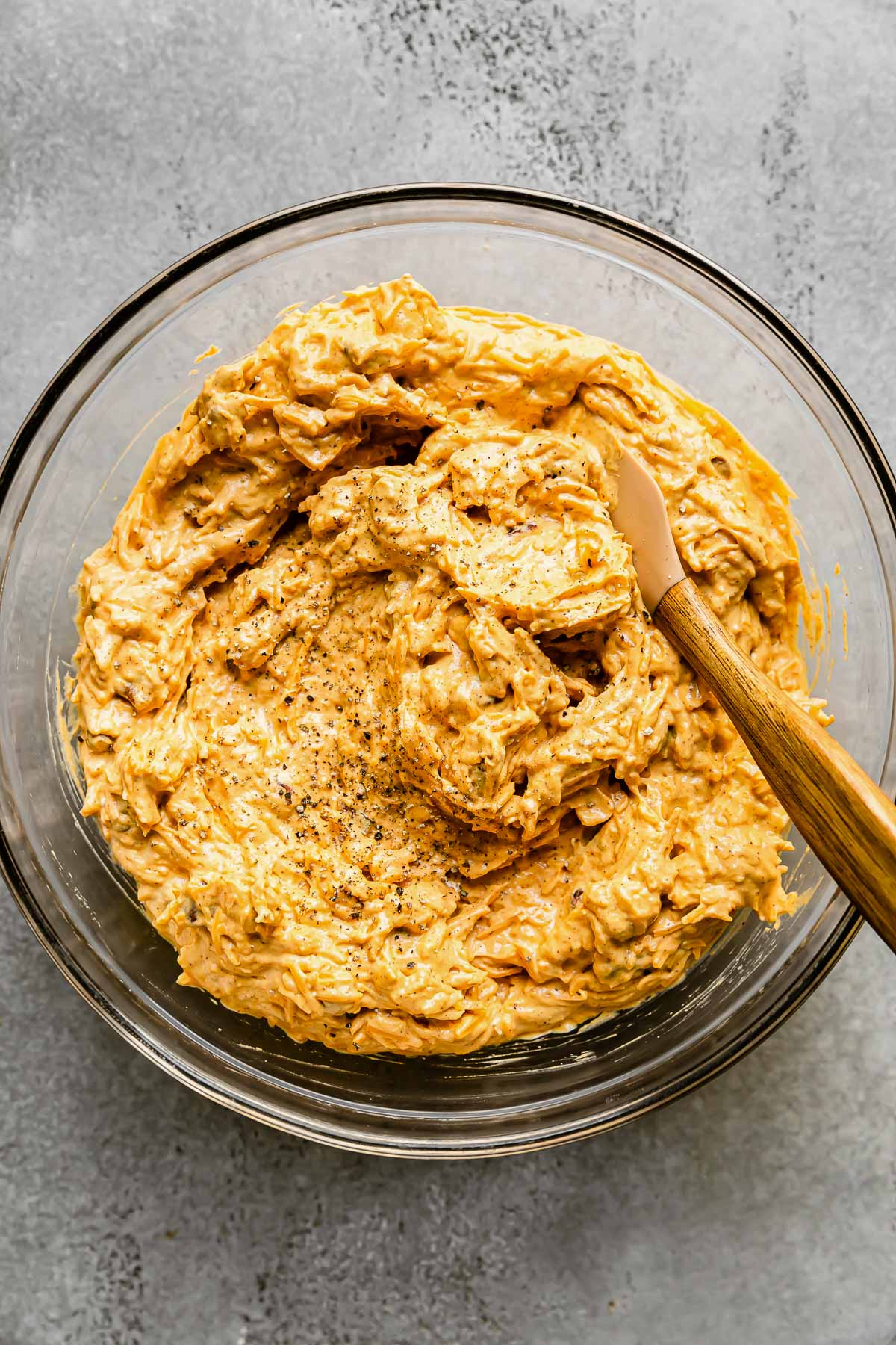 Pumpkin cheeseball mixture fills a large glass mixing bowl that sits atop a gray textured surface. A spatula with a wooden handle rests inside of the bowl for mixing.