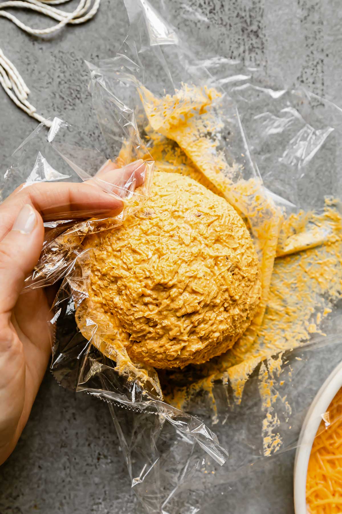 Plastic wrap covers thanksgiving cheese ball mixture that sits atop a gray textured surface. A woman's hands word to shape the cheese ball into a ball shape using the plastic wrap to keep clean. Cut kitchen twine and s small ceramic bowl filled with shredded cheddar cheese surround the formed cheese ball at center.