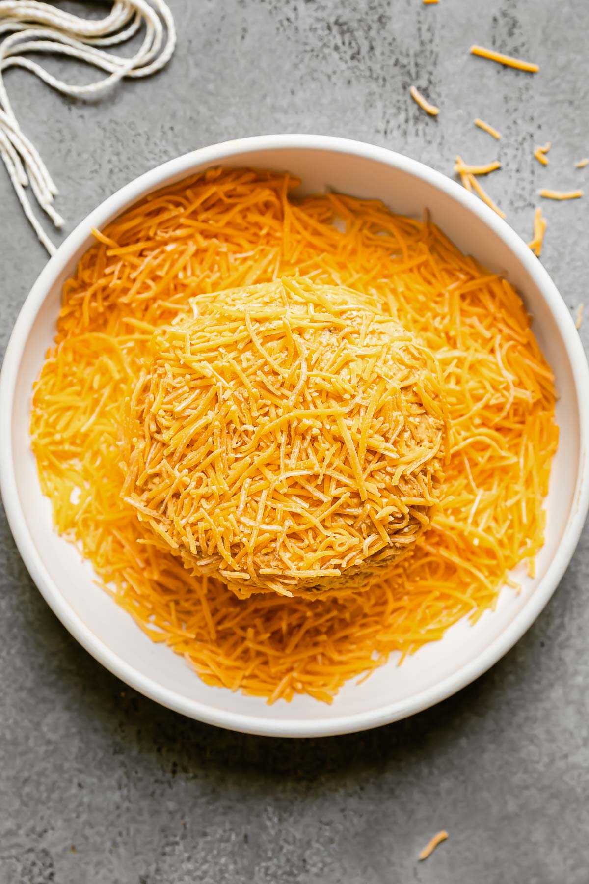 A pumpkin-shaped cheese ball is placed inside a shallow ceramic dish filled with shredded cheddar cheese. The cheese ball gets coated in shredded cheddar cheese and the ceramic dish sits atop a gray textured surface. Loose shredded cheddar cheese and cut strands of kitchen twine surround the dish at center.