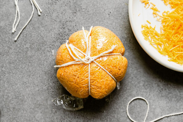 Cut strands of kitchen twine are secured tightly around a formed pumpkin cheese ball wrapped in plastic wrap that sits atop a gray textured surface. The tied kitchen twine creates indentations in the cheese ball to create a pumpkin-shaped cheese ball. The cheese ball is surrounded by a ceramic plate filled with shredded cheddar cheese and other loose cut strands of kitchen twine.