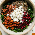 A close up shot of Thanksgiving salad components fill a large gray ceramic bowl: massaged kale, maple spiced roasted sweet potatoes, maple spiced nuts, pomegranate arils, wild rice, and goat cheese. The bowl is surrounded by small containers filled with maple spiced nuts, cinnamon cider vinaigrette, and goat cheese as well as loose fresh sprigs of herbs, a marigold yellow colored linen, and a natural woven placemat with a gold serving spoon and fork resting atop.