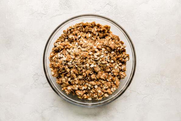 Walnut crumble topping for apple pie bars fills a large glass mixing bowl that sits atop a creamy white textured surface.