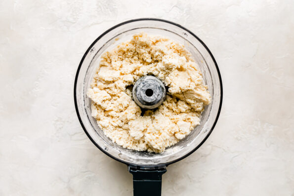 Crust dough for easy apple pie bars fills the bowl of a food processor that sits atop a creamy white textured surface.