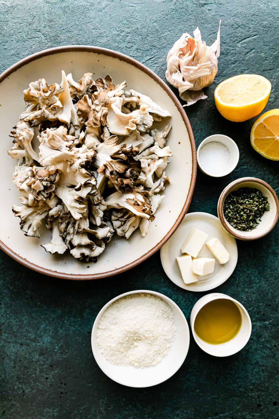 Roasted mushrooms ingredients arranged on a black textured surface: maitake mushrooms, olive oil, kosher salt, unsalted butter, fresh herbs, garlic, lemon, and grated parmesan.