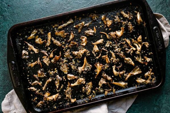 Oven roasted mushrooms are arranged in a single layer atop a metal baking sheet that sits atop a black textured surface. The mushrooms have been tossed with garlic and fresh herbs and have become crispy. A light gray linen napkin rests underneath the baking sheet.
