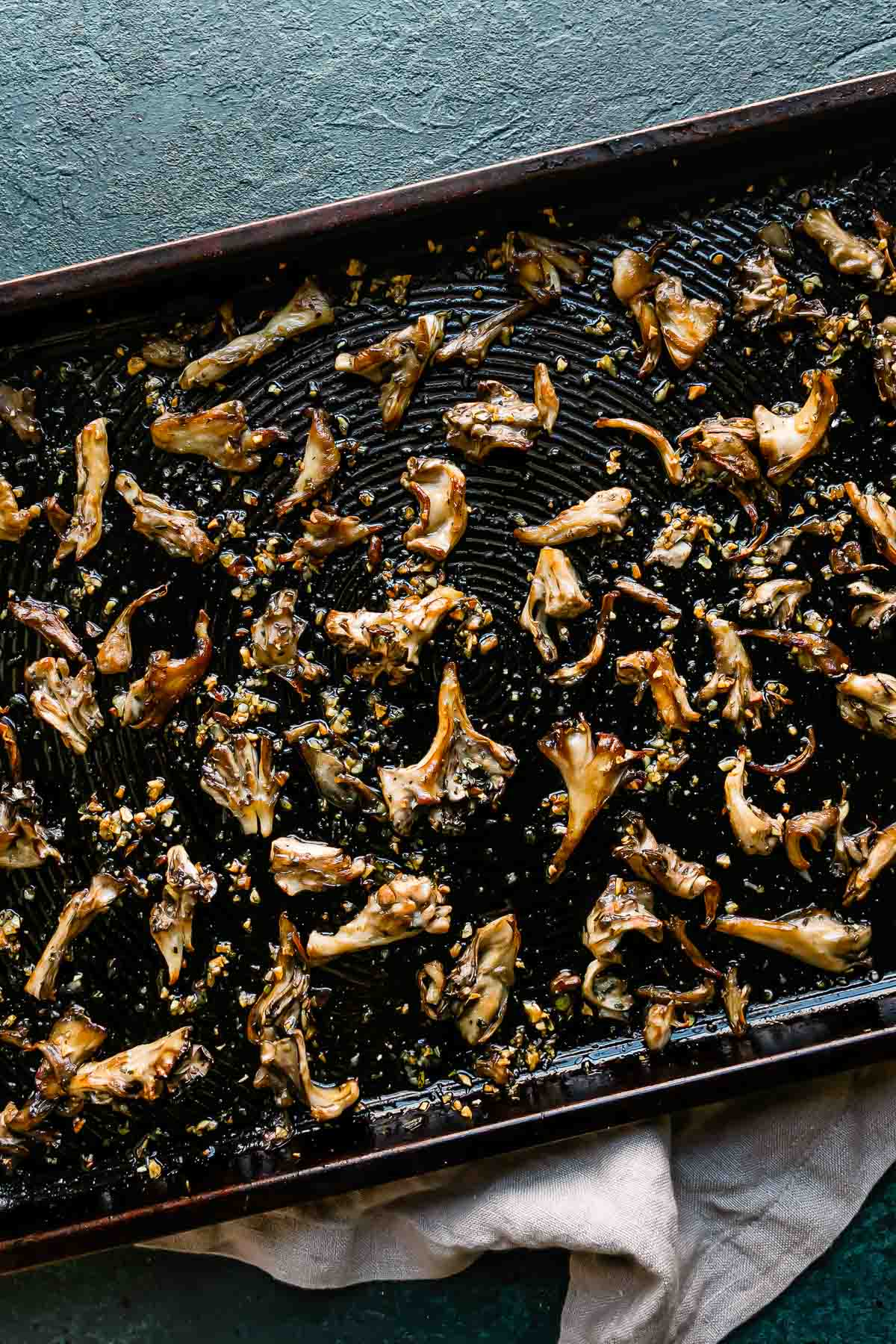 Oven roasted mushrooms are arranged in a single layer atop a metal baking sheet that sits atop a black textured surface. The mushrooms have been tossed with garlic and fresh herbs and have become crispy. A light gray linen napkin rests underneath the baking sheet.