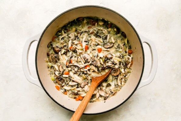 The beginning of easy chicken and wild rice soup fills a large heavy-bottomed pot that sits atop a creamy white textured surface. A wooden spoon rests inside of the pot for stirring.