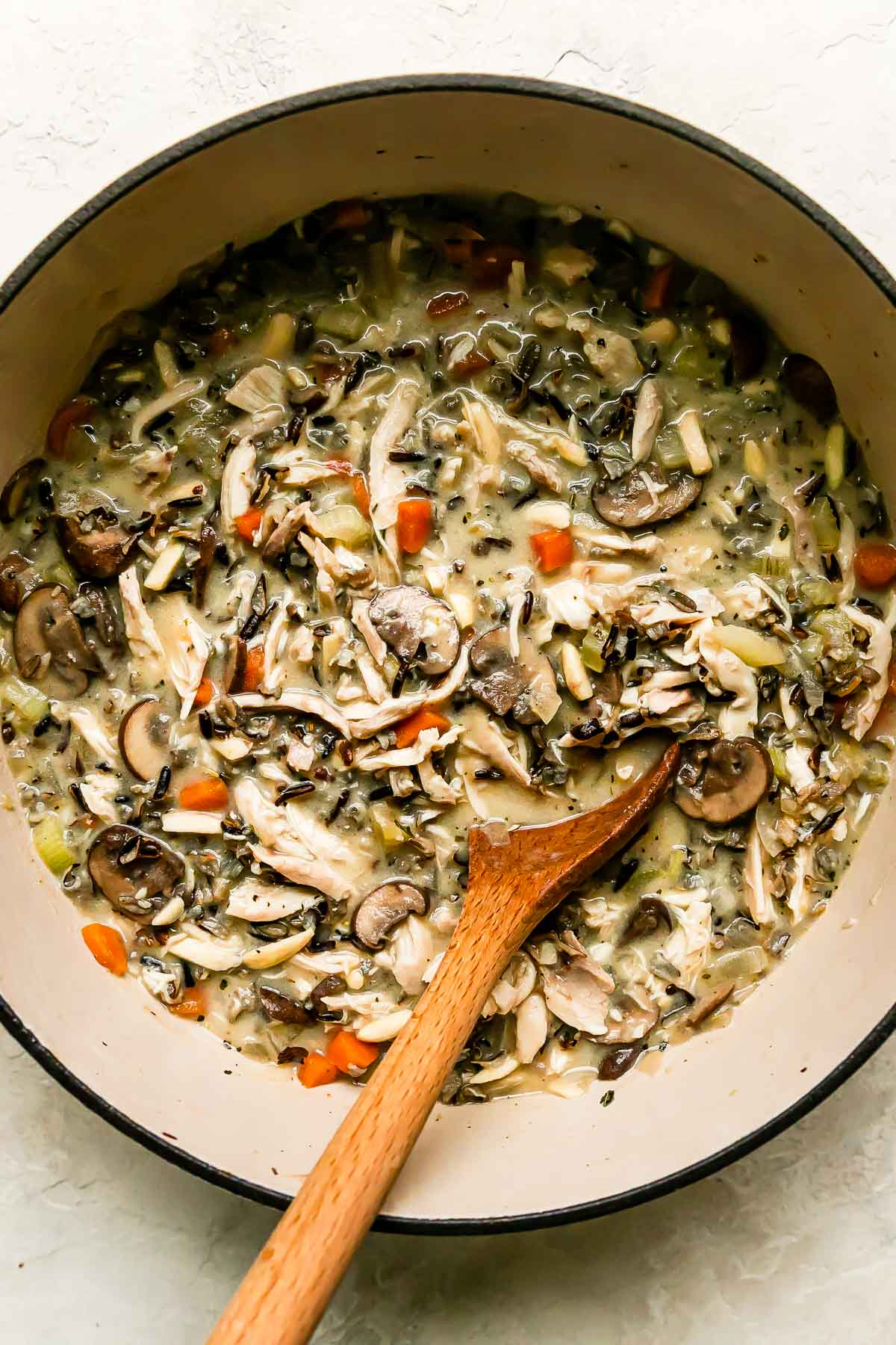 The beginning of easy chicken and wild rice soup fills a large heavy-bottomed pot that sits atop a creamy white textured surface. A wooden spoon rests inside of the pot for stirring.