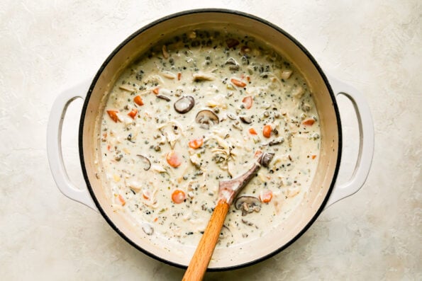 Finished creamy chicken and wild rice soup fills a large heavy-bottomed pot that sits atop a creamy white textured surface. A wooden spoon rests inside of the pot for stirring.