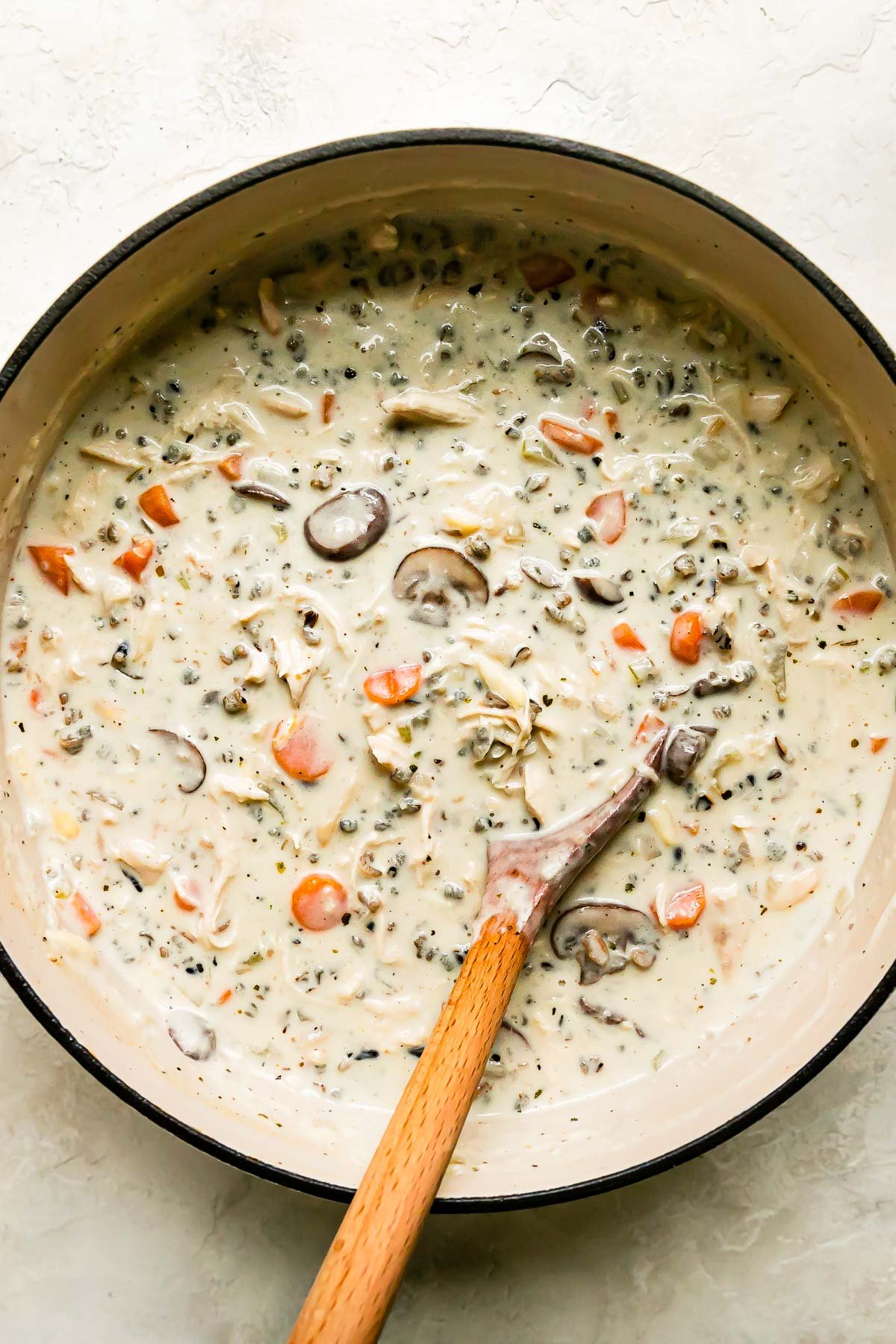 Finished creamy chicken and wild rice soup fills a large heavy-bottomed pot that sits atop a creamy white textured surface. A wooden spoon rests inside of the pot for stirring.