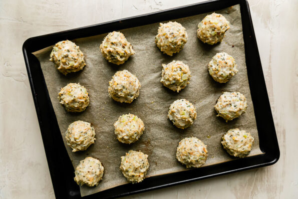16 stuffing meatballs are arranged atop a parchment lined baking sheet that sits atop a creamy white textured surface.