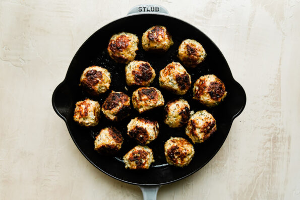16 browned thanksgiving meatballs fill a large gray cast iron skillet that sits atop a creamy white textured surface.