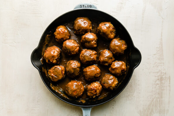 16 thanksgiving meatballs tossed in a homemade gravy fill a large gray Staub cast iron skillet that sits atop a creamy white textured surface.