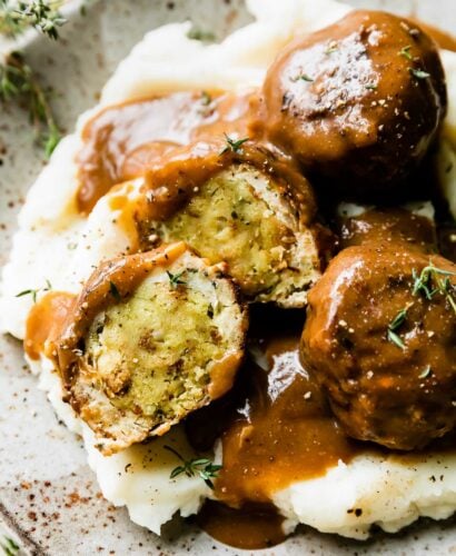 An overhead shot of three thanksgiving meatballs served atop mashed potatoes with gravy on a speckled ceramic plate that sits atop a creamy white textured surface. One of the turkey meatballs have been cut in half to reveal an inside core of stovetop stuffing inside. The meatballs have been drizzled with gravy and garnished with fresh chopped herbs.
