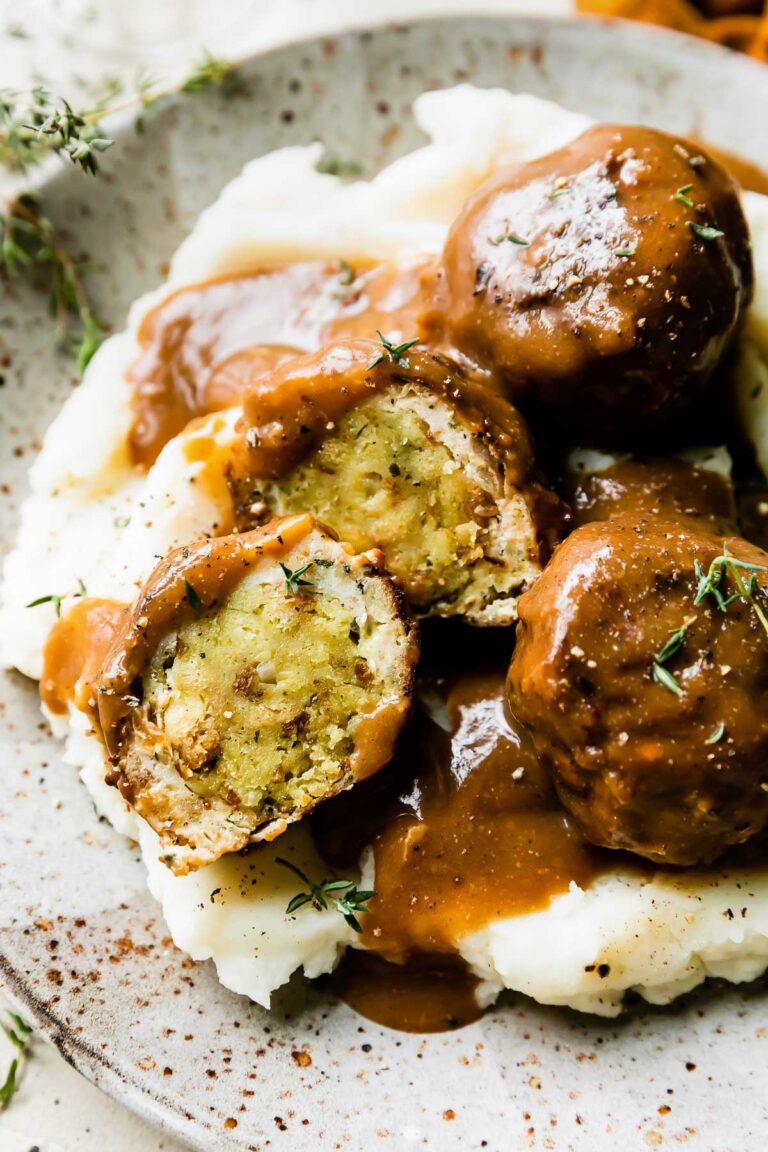 An overhead shot of three thanksgiving meatballs served atop mashed potatoes with gravy on a speckled ceramic plate that sits atop a creamy white textured surface. One of the turkey meatballs have been cut in half to reveal an inside core of stovetop stuffing inside. The meatballs have been drizzled with gravy and garnished with fresh chopped herbs.