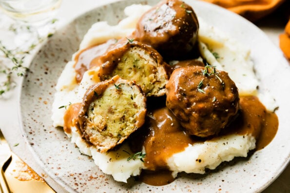 An overhead shot of three thanksgiving meatballs served atop mashed potatoes with gravy on a speckled ceramic plate that sits atop a creamy white textured surface. One of the turkey meatballs have been cut in half to reveal an inside core of stovetop stuffing inside. The meatballs have been drizzled with gravy and garnished with fresh chopped herbs. Gold silverware, fresh sprigs of herbs, a wine glass, and a burnt orange linen napkin surround the plate at center.