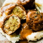 A close up and side angle shot of three thanksgiving meatballs served atop mashed potatoes with gravy on a speckled ceramic plate that sits atop a creamy white textured surface. One of the turkey meatballs have been cut in half to reveal an inside core of stovetop stuffing inside. The meatballs are garnished with fresh chopped herbs and pepper.