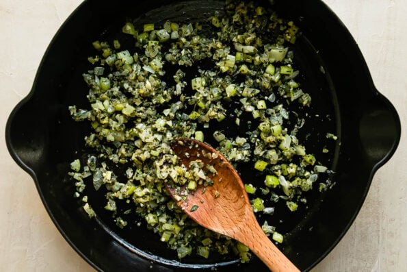 Celery, onion, garlic, and fresh herbs cook inside of a large black cast iron skillet. The skillet sits atop a creamy white textured surface. A wooden spoon rests inside of the skillet for stirring.