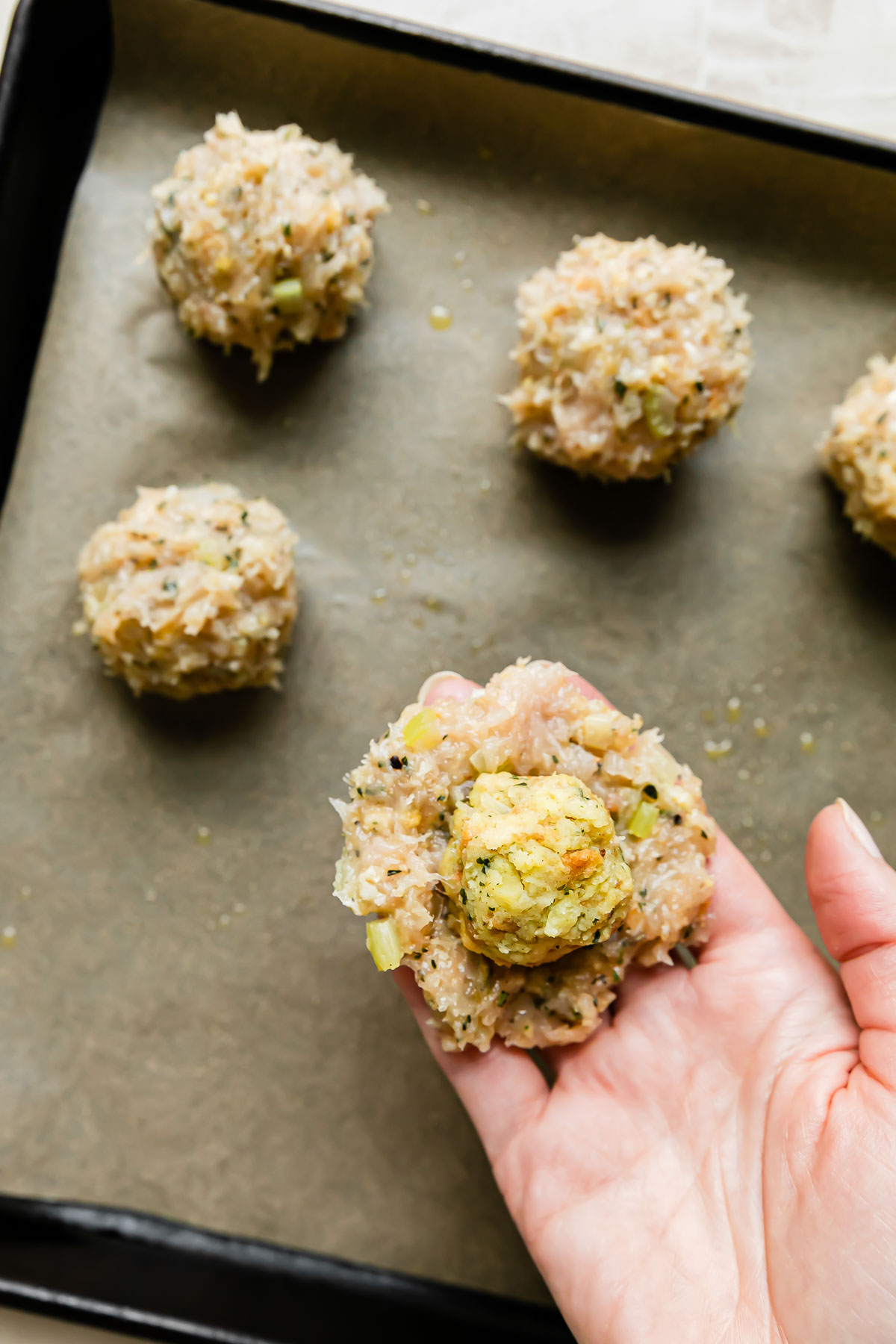 A woman's hand holds a small patty of turkey meatball mixture with a small ball of stovetop stuffing placed in the center of the meatball mixture patty. The patty and stuffing ball are held above a parchment lined baking sheet with stuffed & formed turkey stuffing meatballs arranged atop the parchment paper.