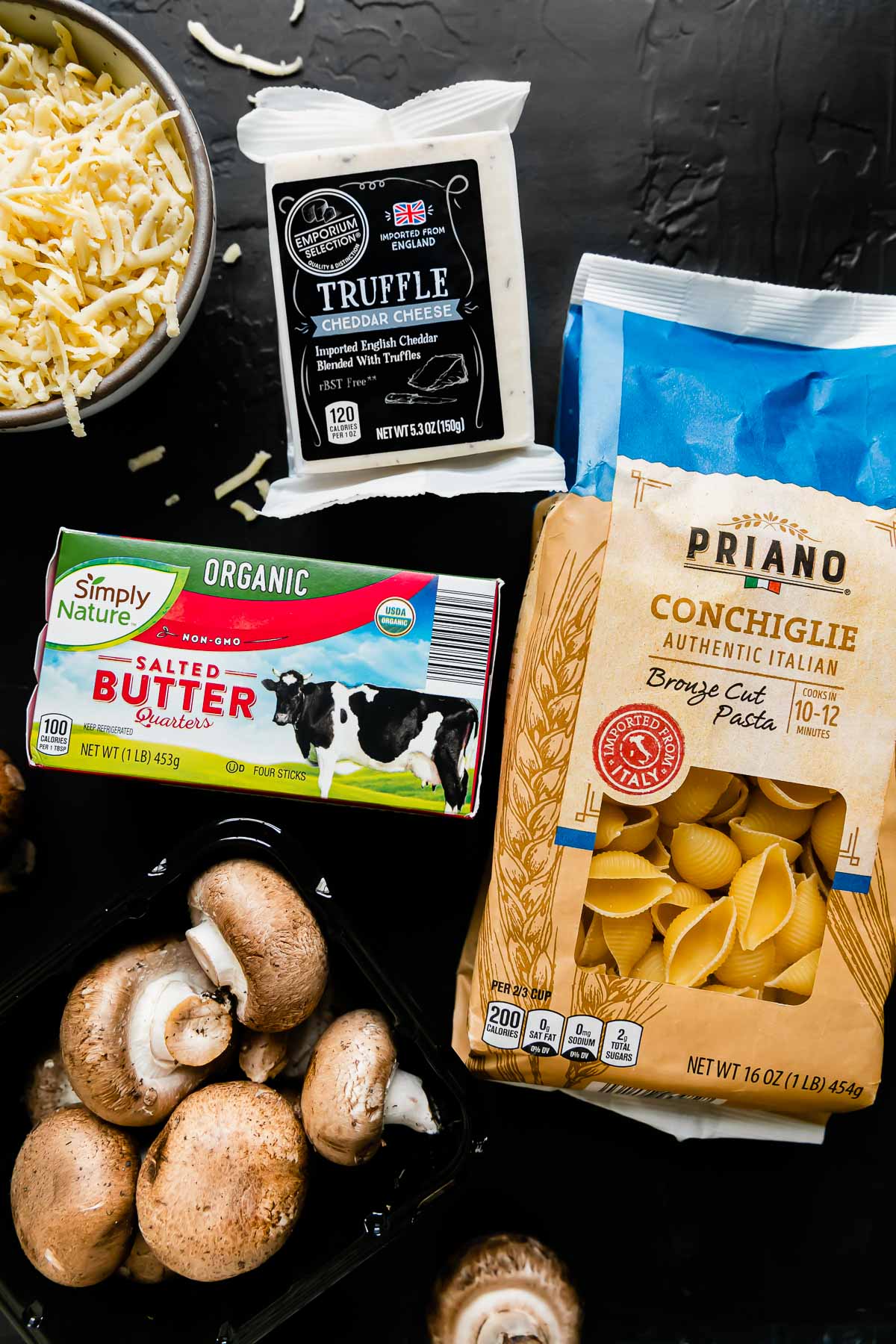Truffle mac and cheese ingredients arranged on a black textured surface: Priano Authentic Italian Bronze Cut Conchiglie Pasta, Simply Nature Organic Salted Butter, cremini mushrooms, and Emporium Selection Premium English Truffle Cheddar Cheese.