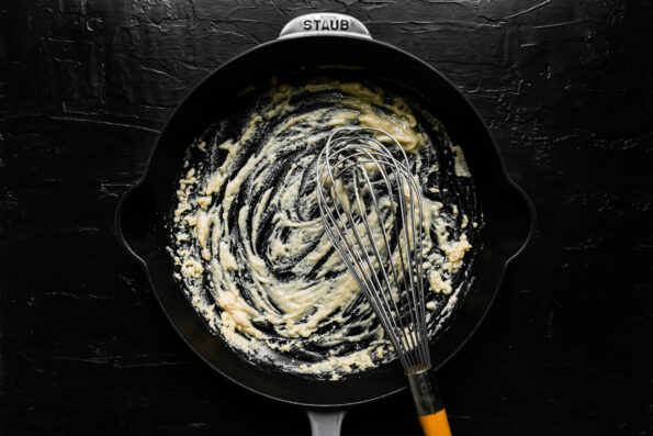 How to make truffled mac and cheese, step 3: Make a roux. A roux is formed inside of a large gray Staub cast iron skillet that sits atop a black textured surface. A metal whisk with a wooden handle rests inside of the pan for stirring.