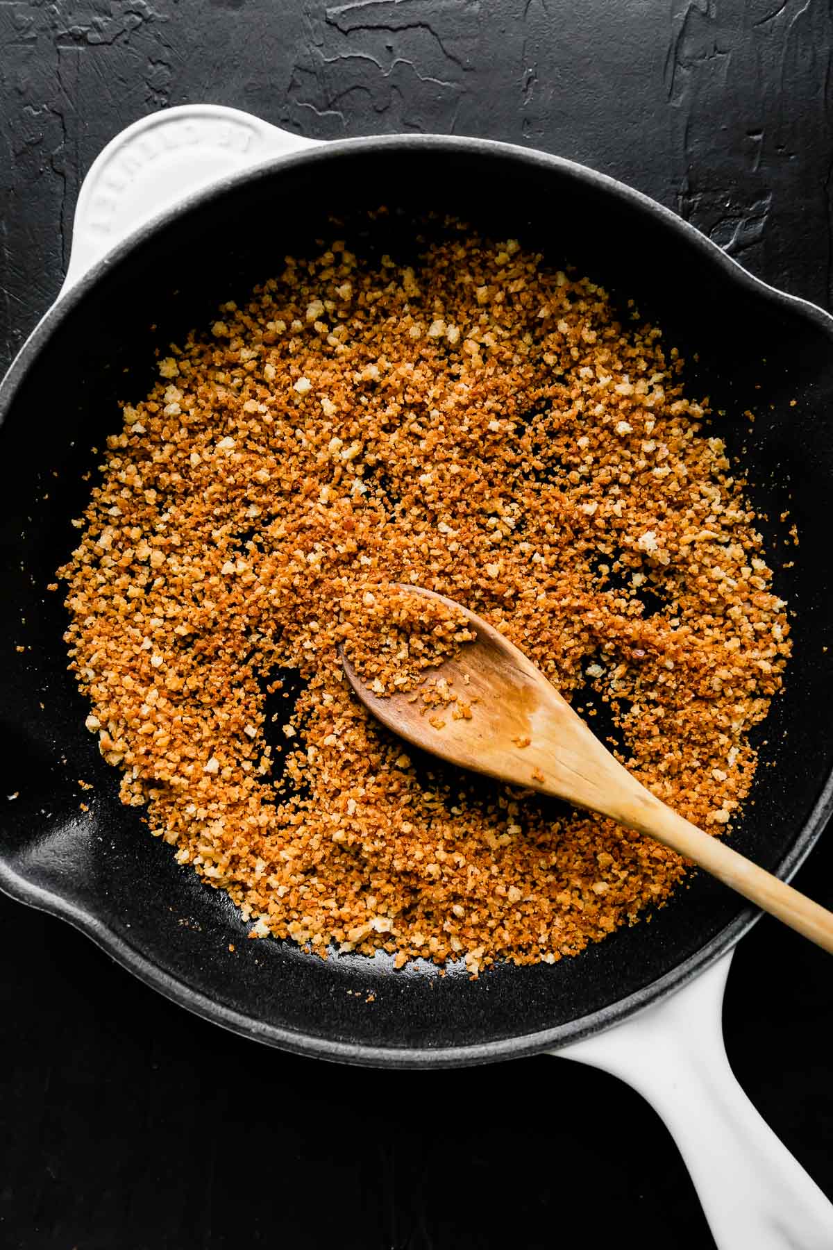 Toasted garlic breadcrumbs fills a small white Le Creuset cast iron pan that sits atop a black textured surface. A wooden spoon rests inside of the pan for stirring.