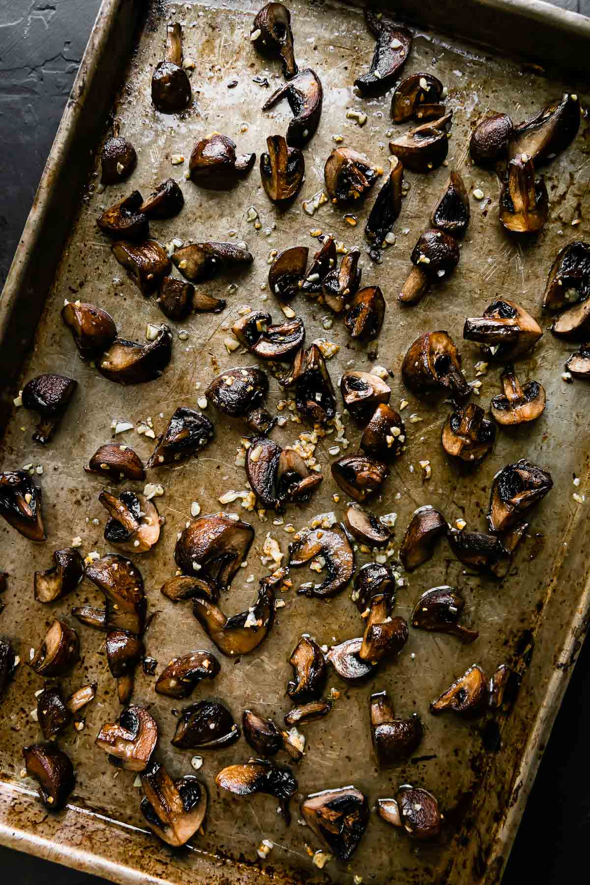 Crispy roasted mushrooms are arranged atop a metal baking sheet. The baking sheet sits atop a black textured surface.