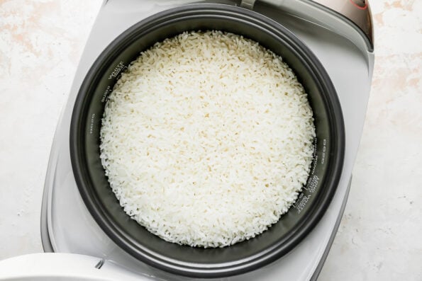 An overhead shot of perfectly cooked white rice for poke bowls rests inside of an open Zojirushi rice cooker. The rice cooker sits atop a creamy white textured surface.