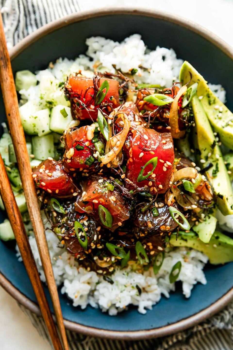 An ahi tuna poke bowl is assembled inside of a large blue ceramic bowl. The bowl sits atop a creamy white textured surface and a pair of wooden chop sticks rests atop the bowl. A blue and white linen napkin is tucked underneath the bowl.