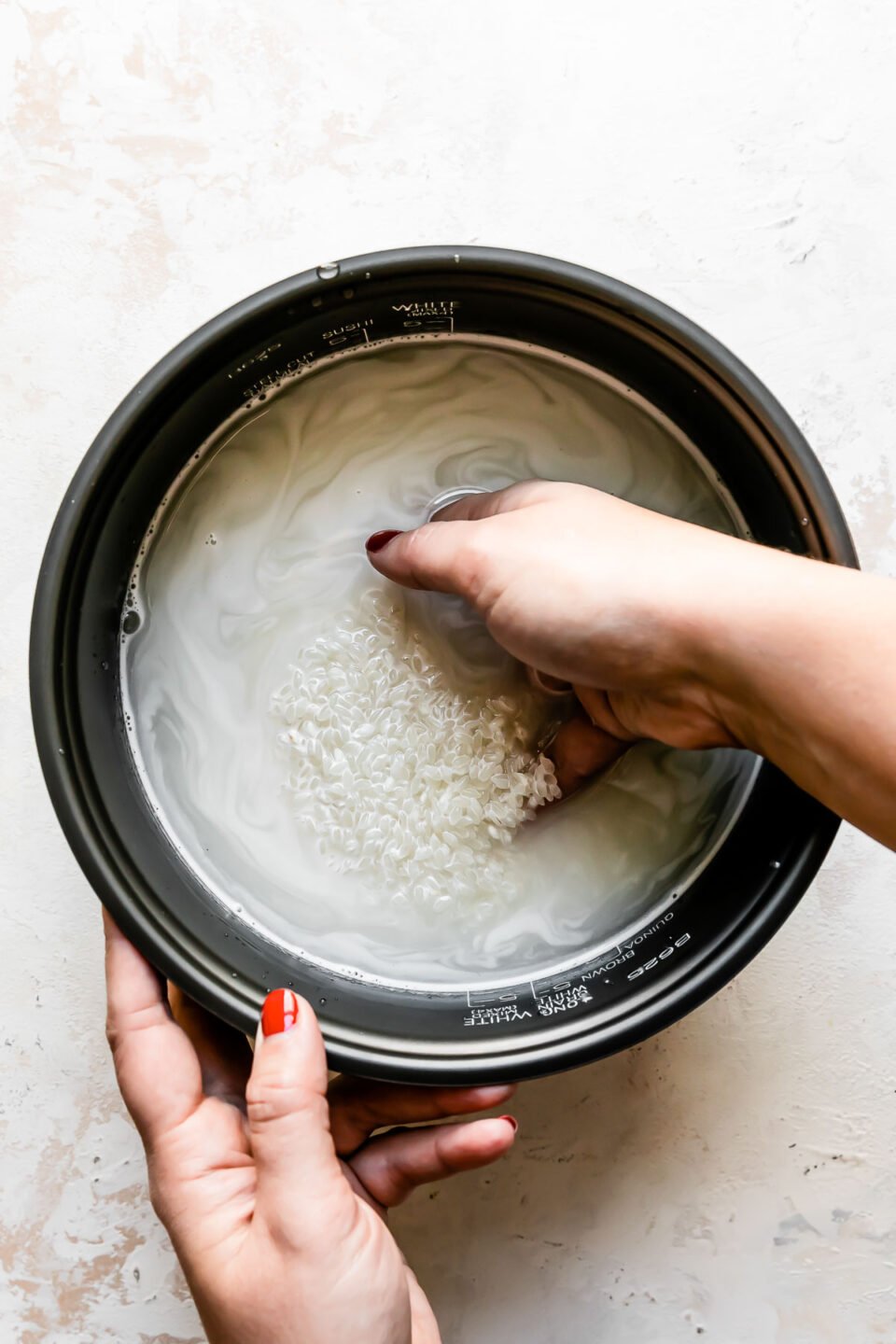 A woman's hand works to rinse white rice in water inside of the inner cooking pan of a Zojirushi rice cooker. The pan sits atop a creamy white textured surface.