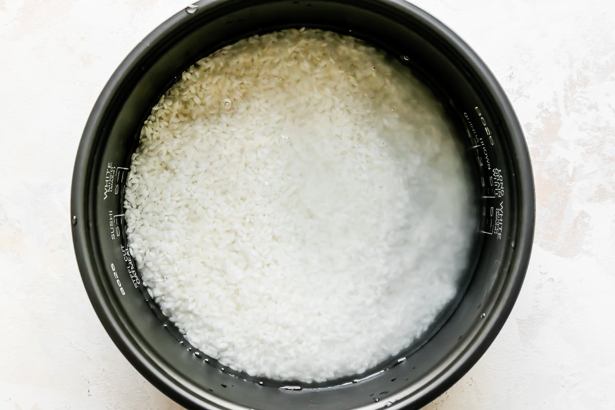 Rinsed white rice in water fills the inner cooking pan of a Zojirushi rice cooker. The pan sits atop a creamy white textured surface.
