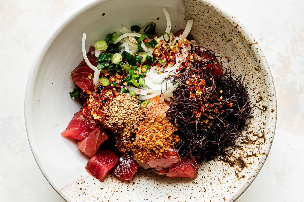 Ahi poke bowl components are arranged in a large ceramic bowl that sits atop a creamy white textured surface: ahi tuna, sweet onion, green onions, ogo or limu, shoyu, toasted sesame oil, Hawaiian sea salt, Inamona, toasted sesame seeds, and crushed red pepper.